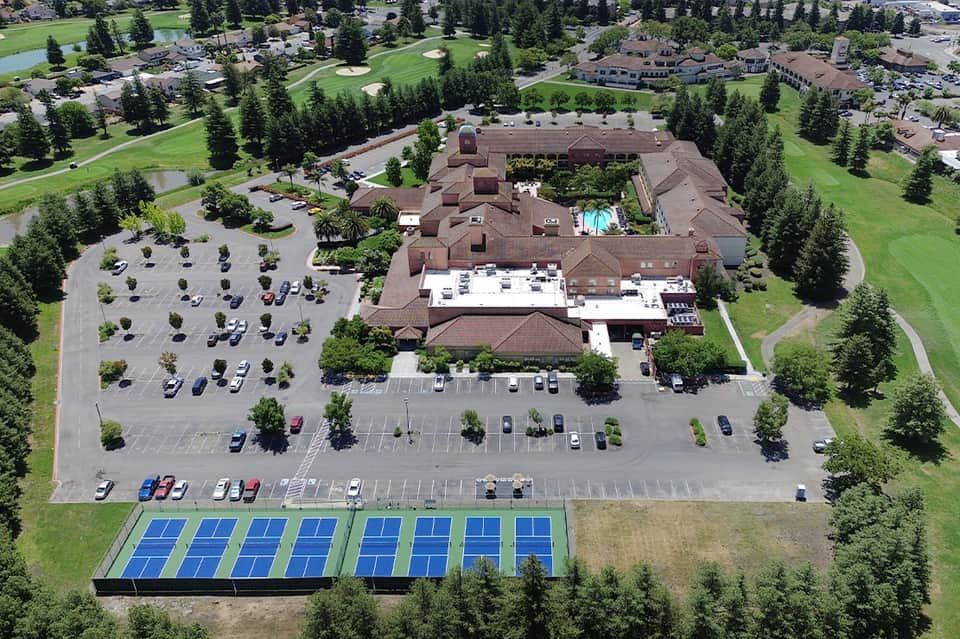 Aerial view of resort property with parking lot, pool, tennis courts, and surrounding green landscape