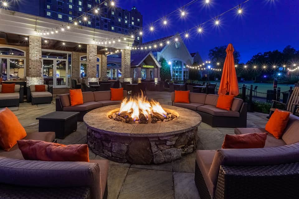 Outdoor fire pit seating area at night with brown sofas, orange pillows, and string lights