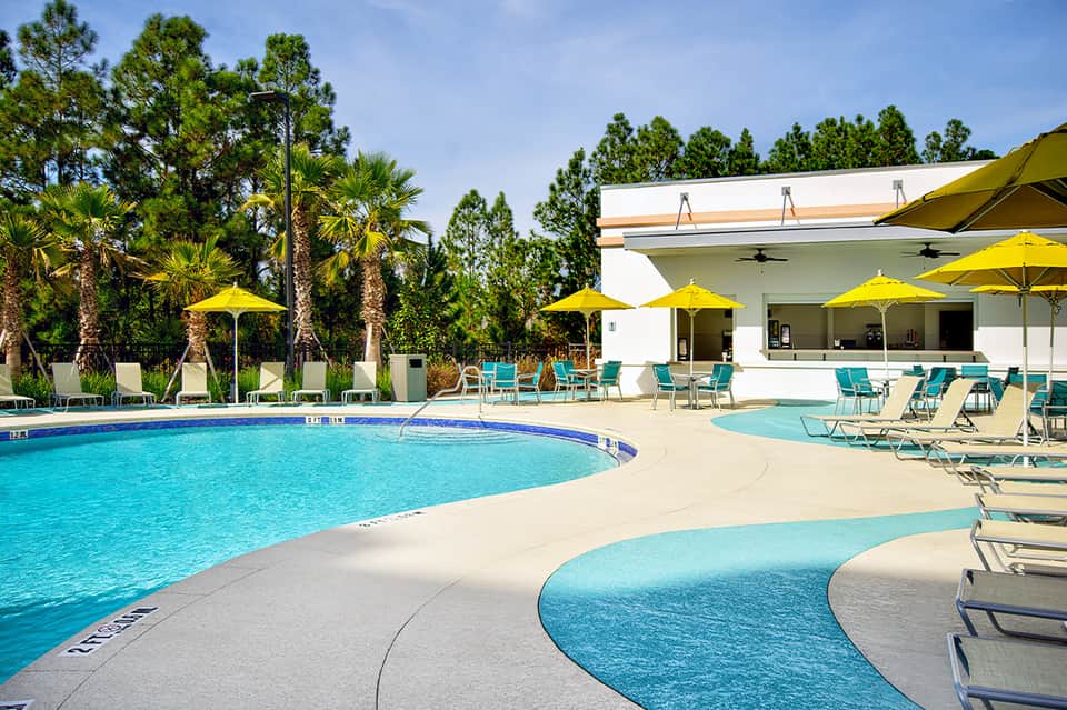 Resort pool with curved design, yellow umbrellas, palm trees, and white clubhouse building