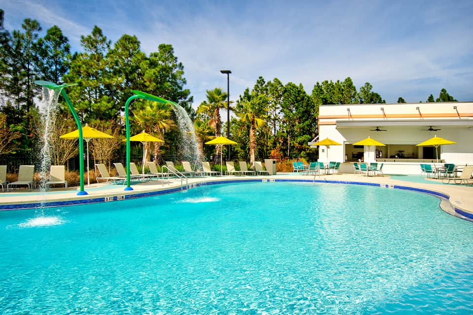 Bright aquamarine pool with splash pad features, green arch, yellow umbrellas, and white pavilion building
