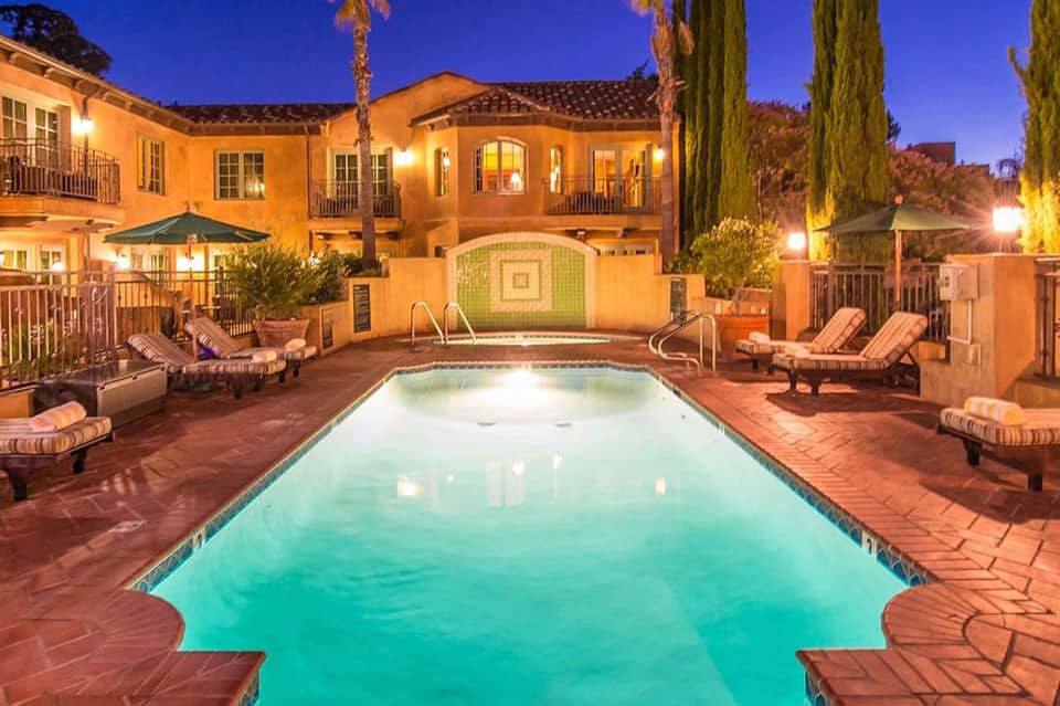 Illuminated courtyard pool at dusk with turquoise water, lounge chairs, palm trees, and Spanish-style architecture