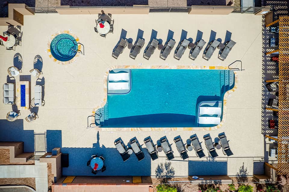 Aerial view of resort pool deck with blue pool, lounge chairs, hot tub, and beige deck area