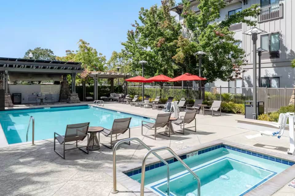 Rectangular pool with lounge chairs, red umbrellas, and pergola-covered patio area
