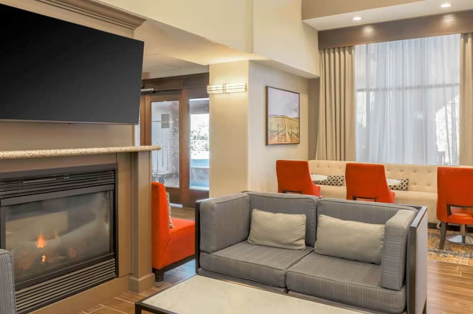 Modern hotel lounge with fireplace, orange accent chairs, gray sofa, flat-screen TV, and floor-to-ceiling windows