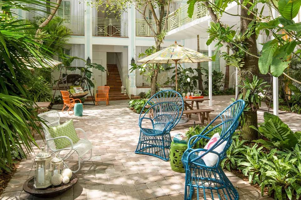 Courtyard with white colonial building, tropical plants, blue wicker chairs, wooden table with umbrella, and orange accent seating