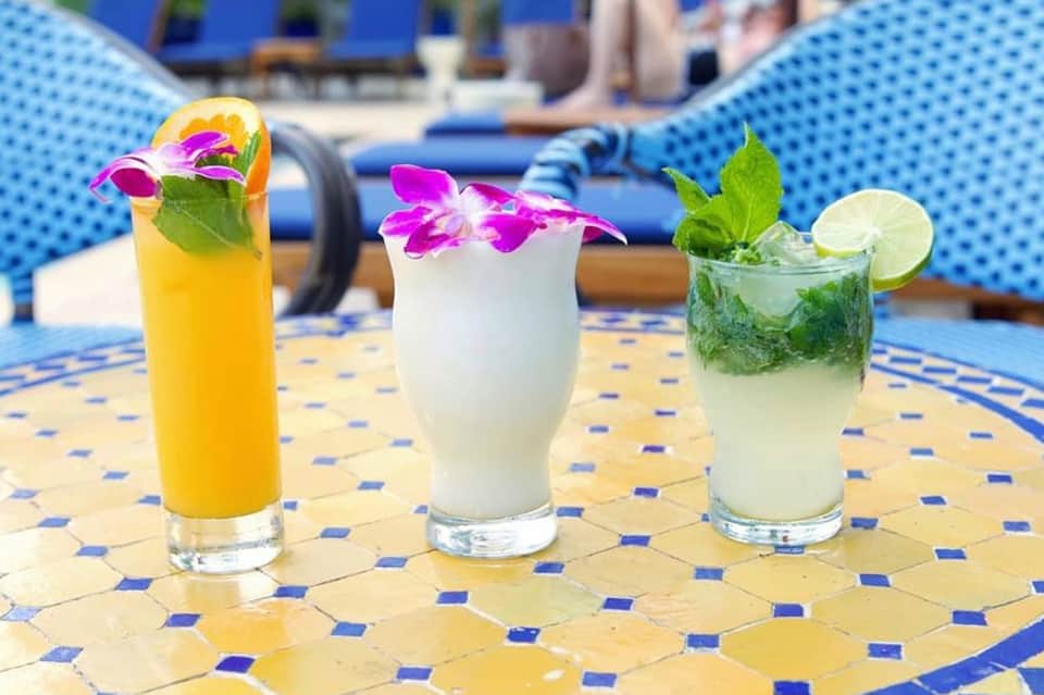 Three colorful cocktails with flowers on mosaic poolside table, blue loungers behind