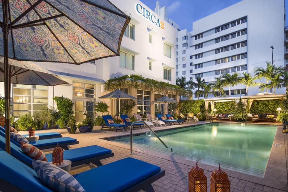 Hotel pool patio at dusk with blue loungers, umbrellas, lit building, and landscaping