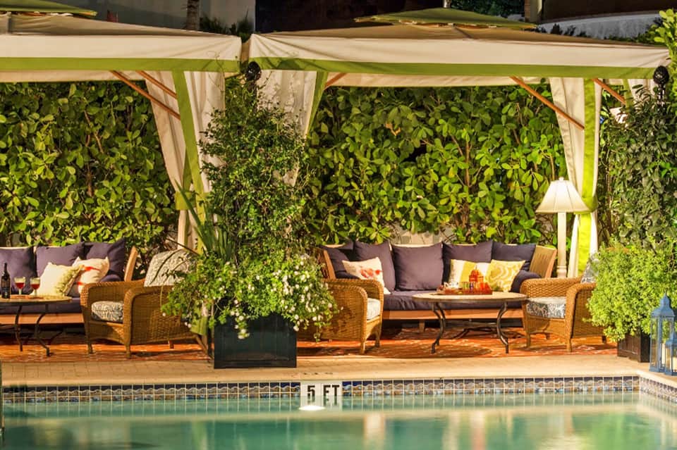 Poolside seating area with woven furniture, cream pergola, colorful cushions, and green foliage