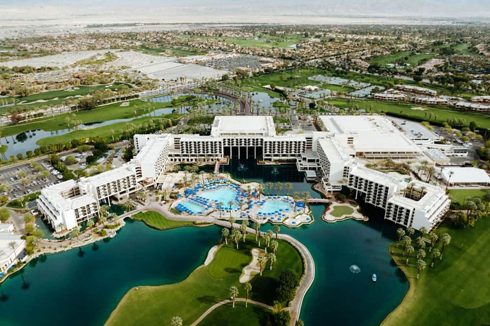 Aerial view of large resort with multiple white buildings, pools, surrounding golf courses and water features