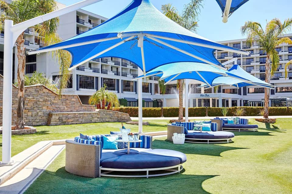 Resort lawn area with blue sail shade structures, round lounge beds with cushions, and palm trees