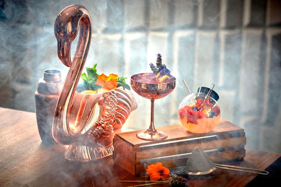 Artistic cocktail display with pink glassware, flowers, and smoke effect on wooden surface
