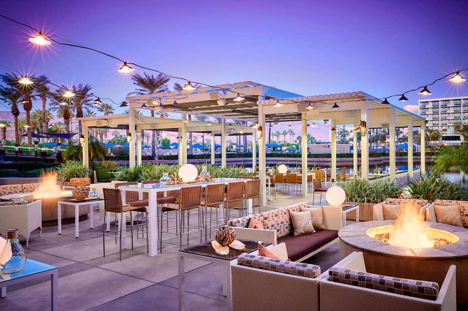 Modern waterfront restaurant and bar at dusk with fire pits and lounge seating under pergolas