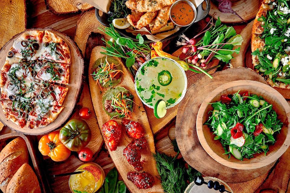 Overhead spread of colorful gourmet food including pizza, grilled proteins, fresh salads, and bread