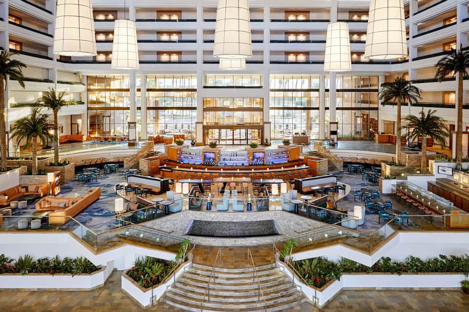 Grand multi-story hotel atrium with circular lounge area, palm trees, and expansive windows