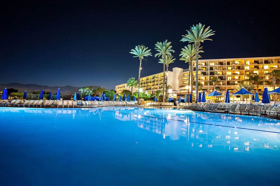Illuminated resort pool at night with blue umbrellas, palm trees, and lit hotel building