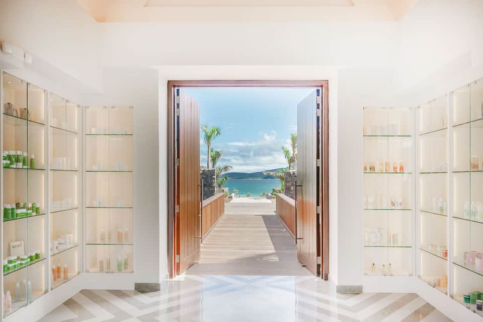 Luxury spa corridor with glass shelving displaying products, leading to ocean view terrace with palm trees