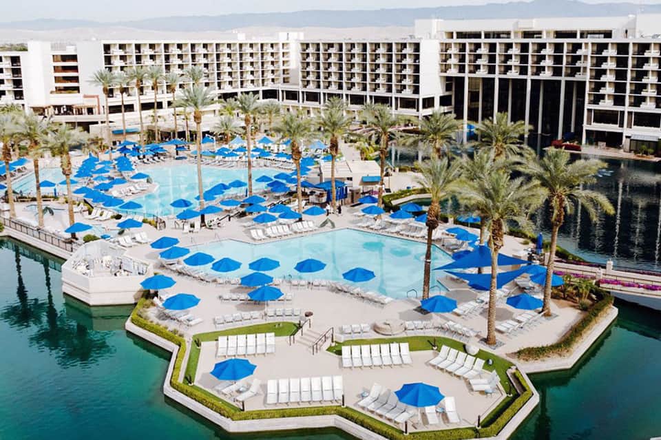 Resort pool area with blue umbrellas, palm trees, and modern white buildings