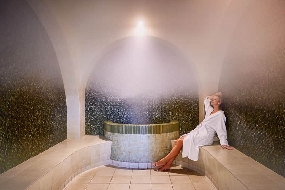 Woman in white robe relaxing in luxurious spa steam room with mosaic tile walls and spa tub