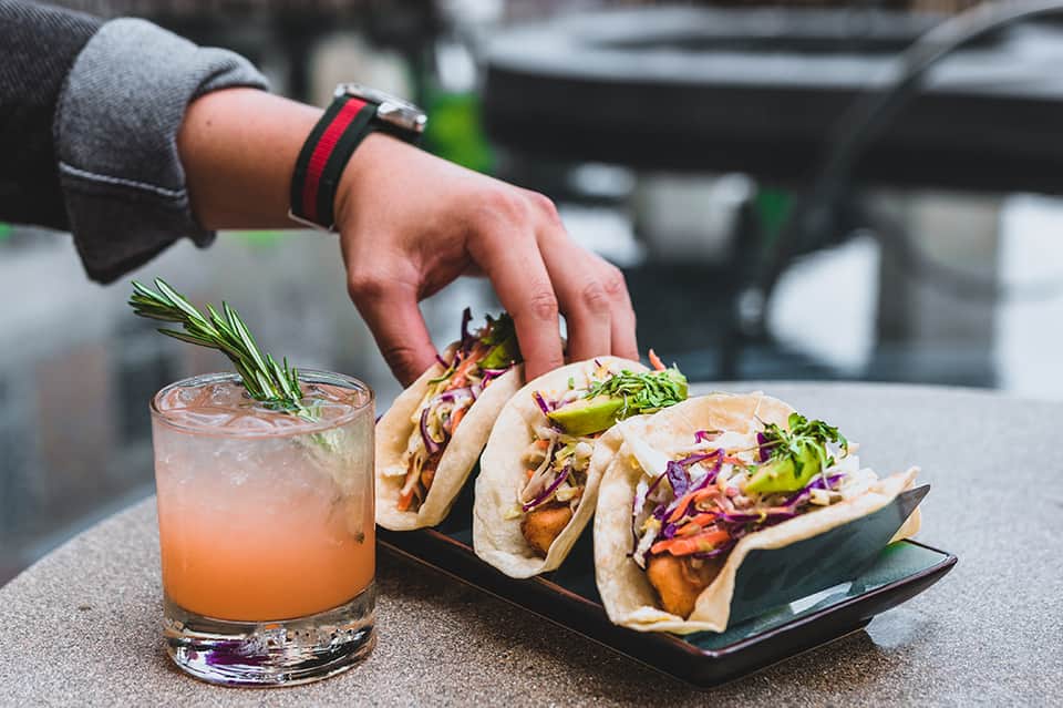 Hand holding gourmet tacos with drink, featuring purple cabbage slaw and fresh herbs