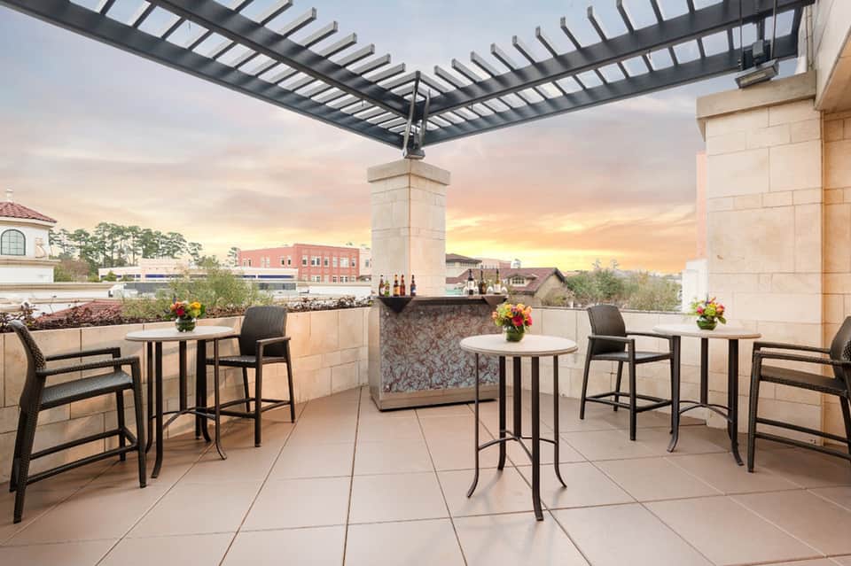 Rooftop bar terrace at dusk with high-top tables, bar counter, pergola, and city skyline views