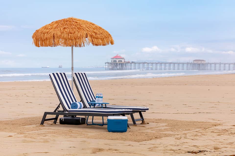 Two striped beach lounge chairs under thatched umbrella on sandy beach with pier and ocean in background