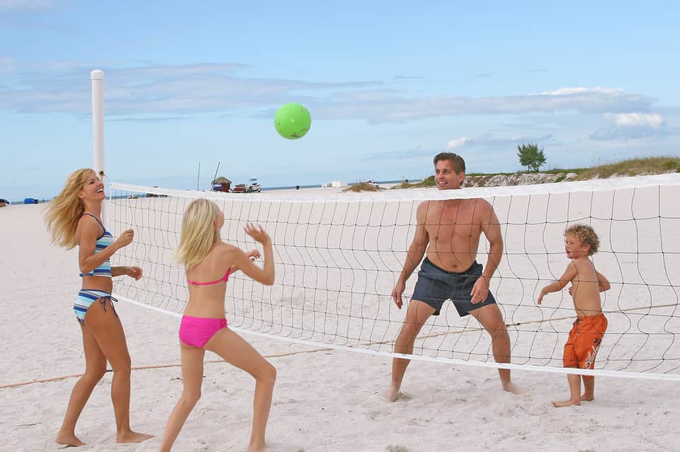 Family playing volleyball on beach with net, ocean and shoreline visible in background
