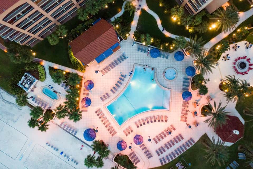 Aerial view of illuminated resort pool complex with umbrellas and palm trees at dusk