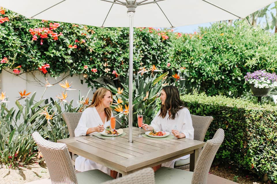 Two women dining at outdoor garden table under umbrella with flowering plants and green hedges