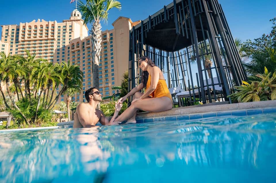Couple in pool at resort with modern black structure, high-rise hotel, and palm trees
