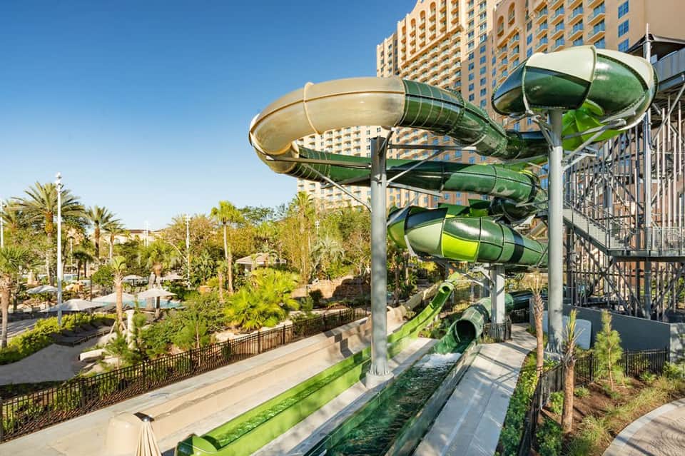 Water park with green spiral slides, palm trees, high-rise hotel building, and shallow recreational pool area