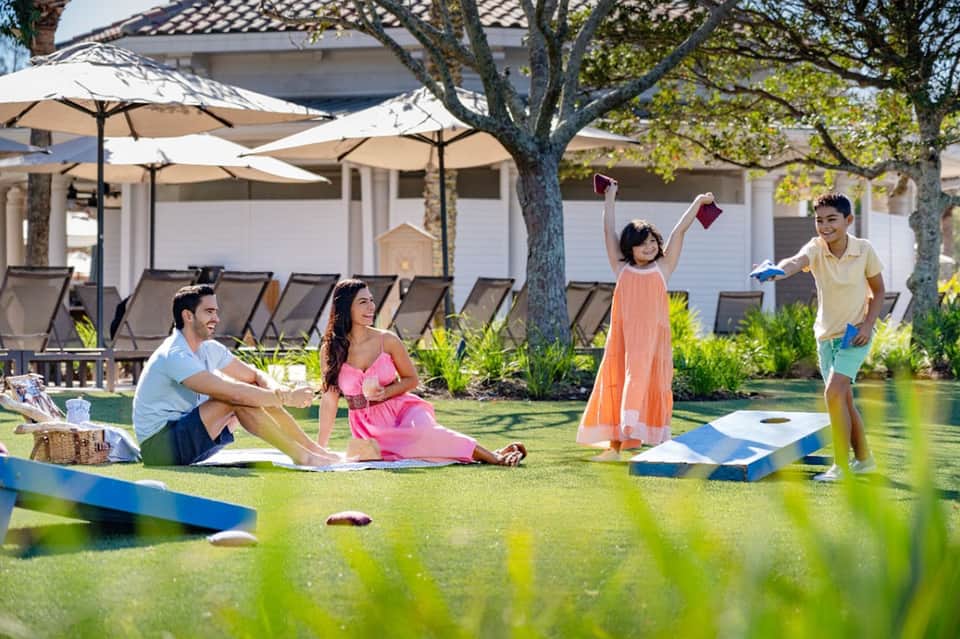 Family playing cornhole on lawn with pool, umbrellas, and beach cabanas in background