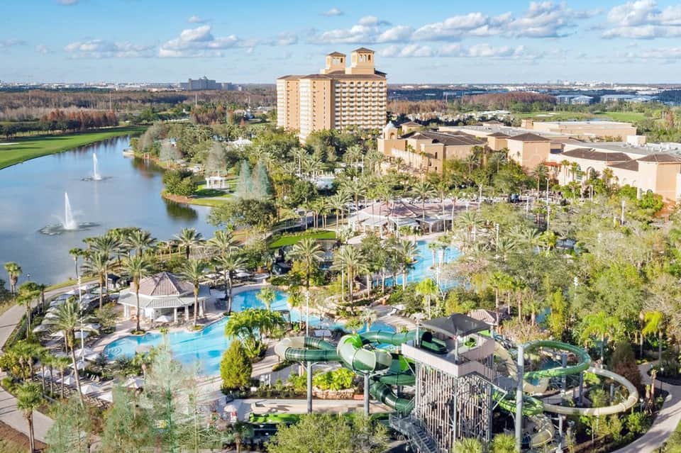 Aerial view of sprawling resort with multiple pools, water slides, hotel towers, and lake