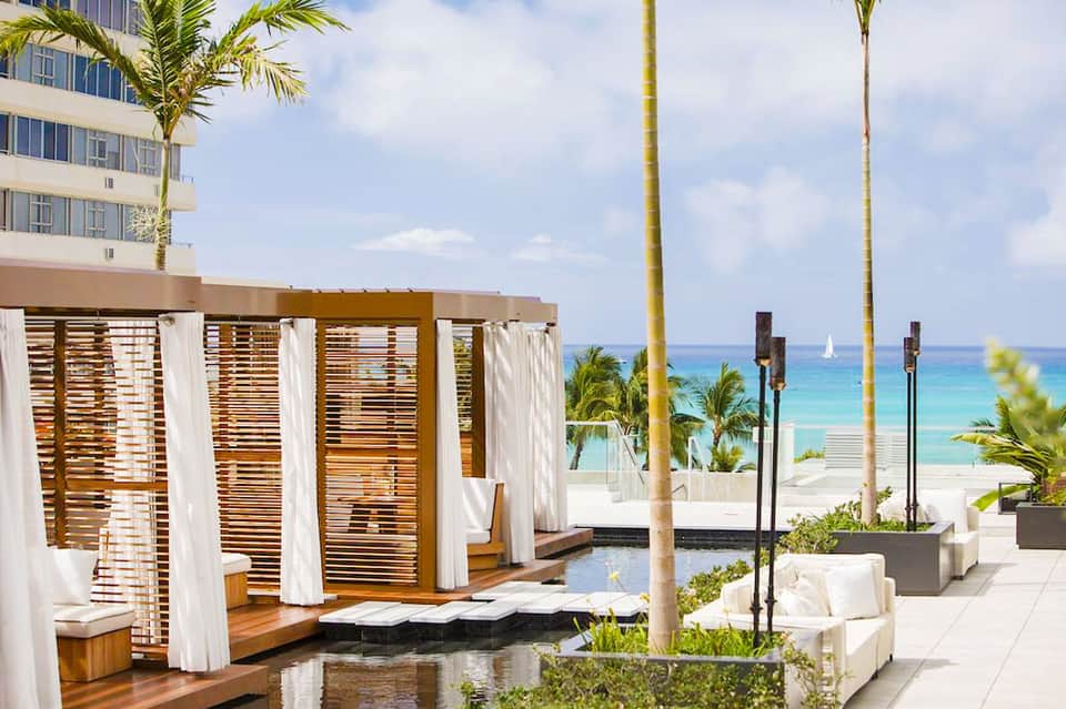 Luxury beachfront resort deck with wooden cabanas, white curtains, and ocean view beyond palm trees