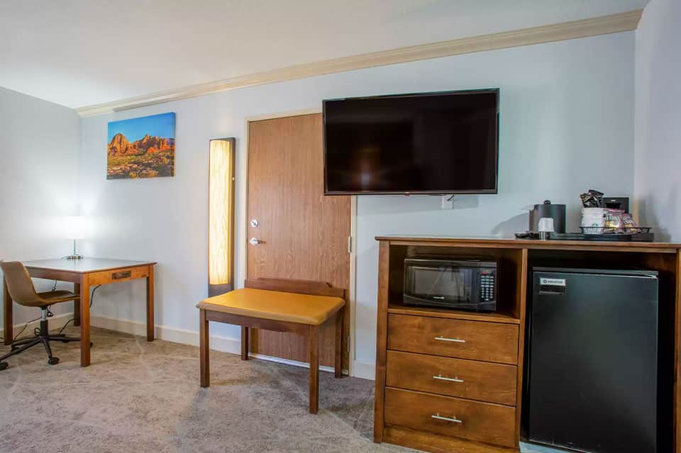 Hotel room interior with wooden furniture, TV, desk, microwave, and refrigerator in neutral tones