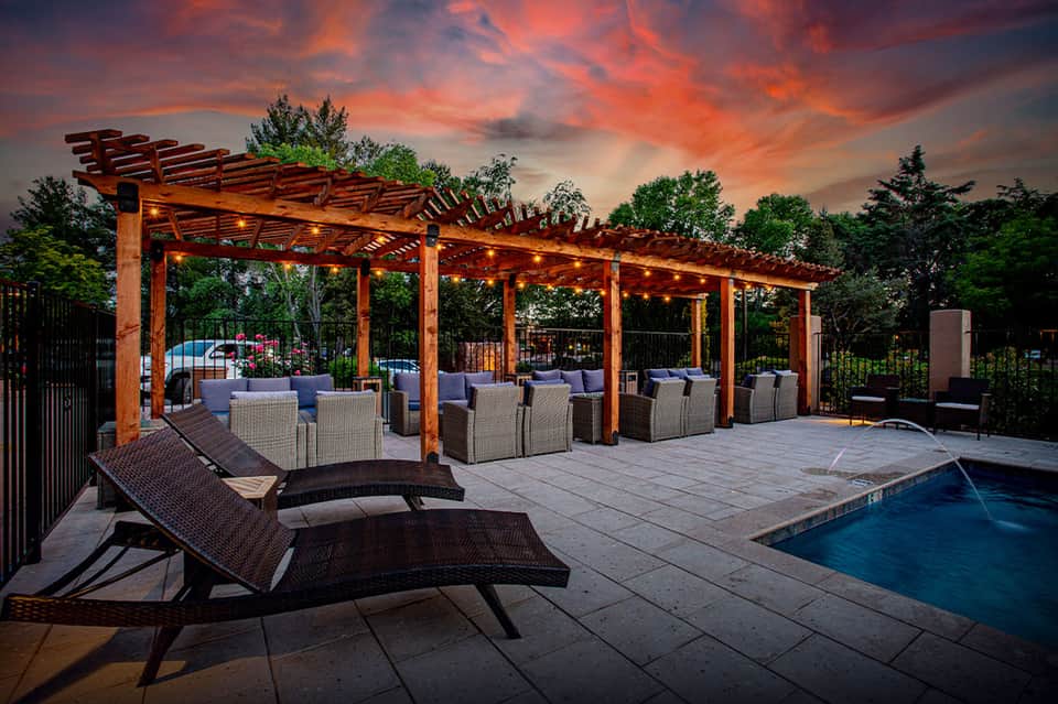Resort pool deck at dusk with wooden pergola, seating area, lounge chairs, and glowing lights