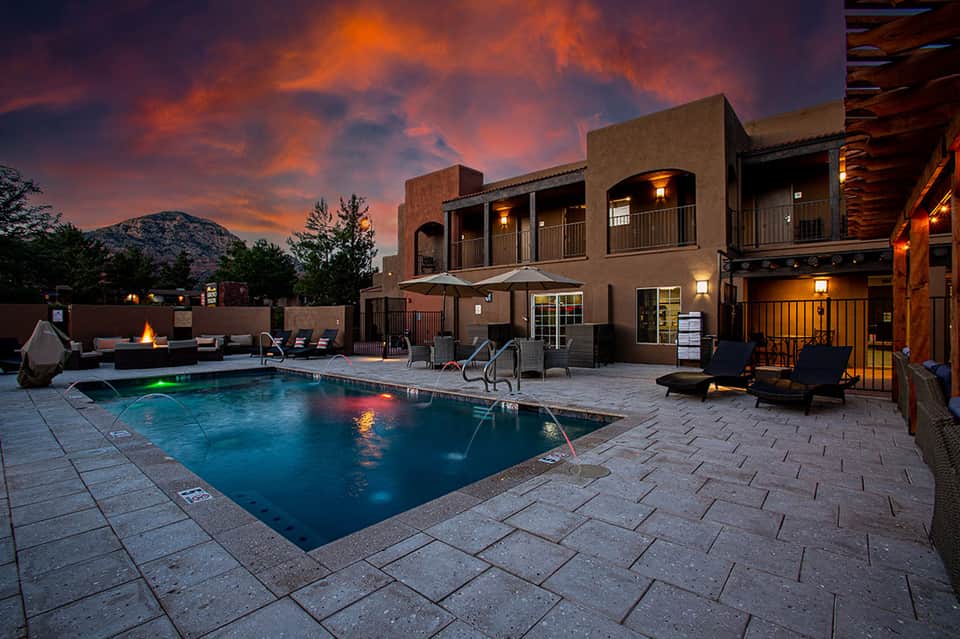 Illuminated resort pool at dusk with fire pit, lounge chairs, and Southwestern-style building against sunset sky