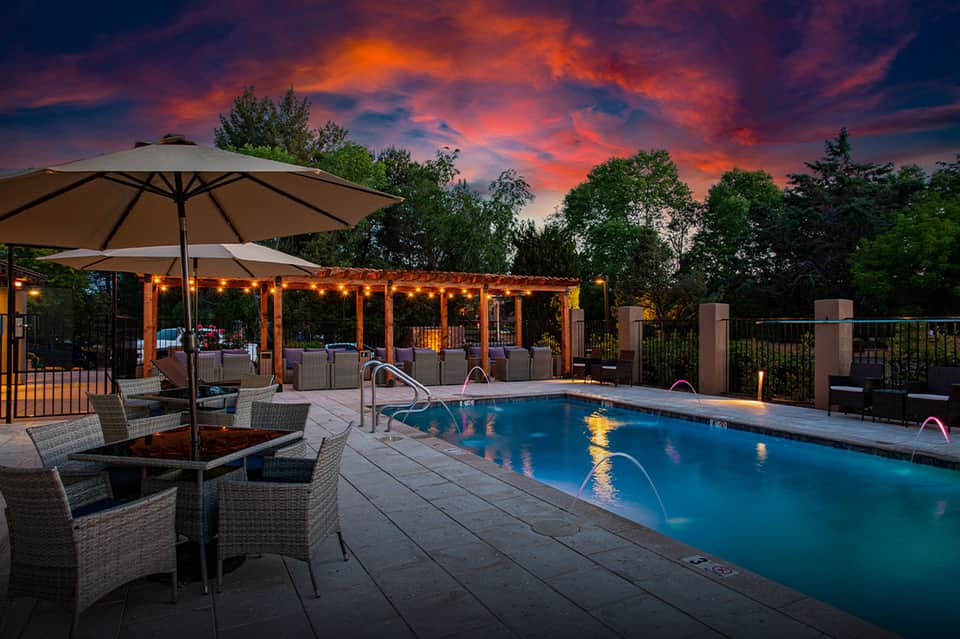 Illuminated poolside area at dusk with umbrellas, lounge chairs, and pergola seating