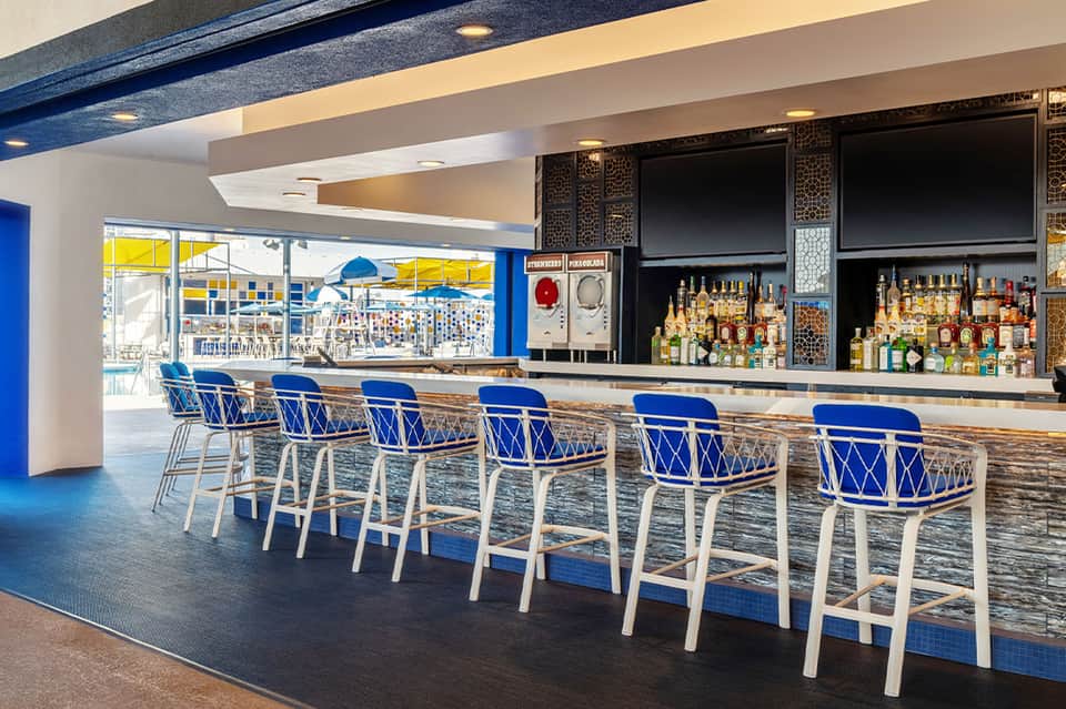 Modern beachfront bar with blue barstool seating, ocean view windows, and fully stocked liquor shelves