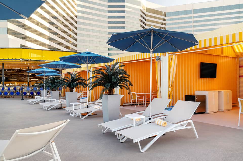 Rooftop lounge with blue umbrellas, white loungers, and striped orange container structures