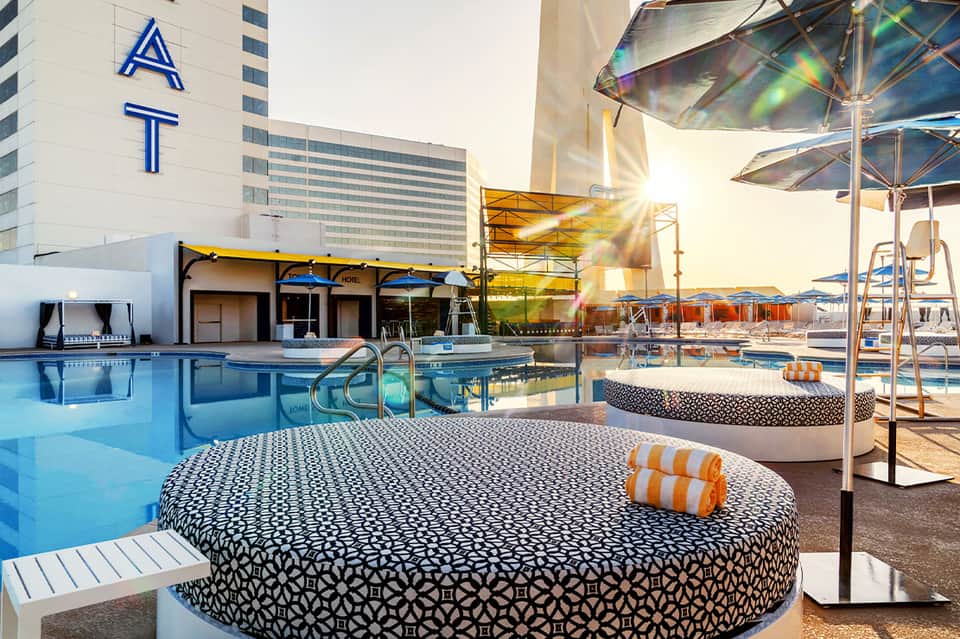 Resort pool deck with modern loungers, umbrellas, water features, and hotel building