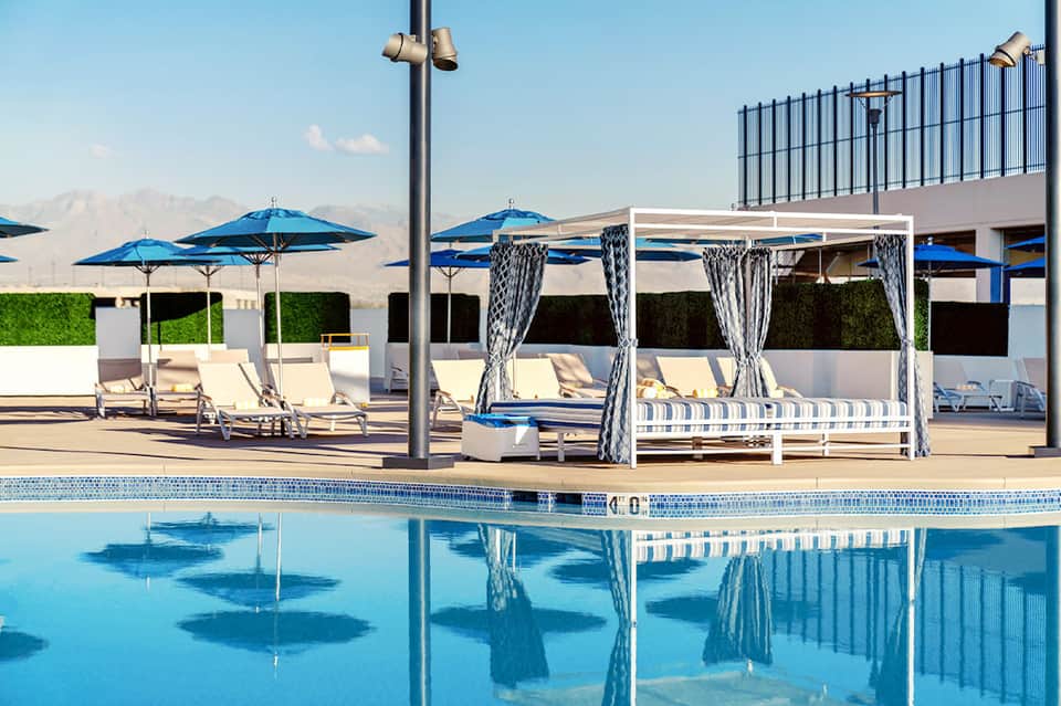 Resort poolside with blue umbrellas, white cabanas with curtains, lounge chairs, and mountain backdrop