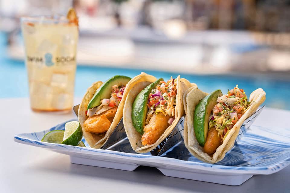 Three fish tacos with avocado and coleslaw on decorative plate by pool area