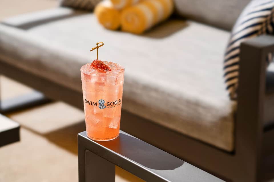 Colorful cocktail with 'Swim & Social' branding on poolside table with lounge seating
