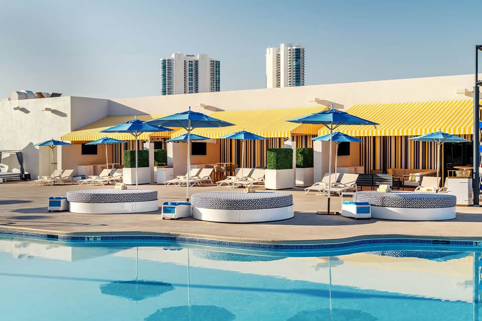 Rooftop pool deck with blue umbrellas, cabanas, yellow striped canopy, and city skyline