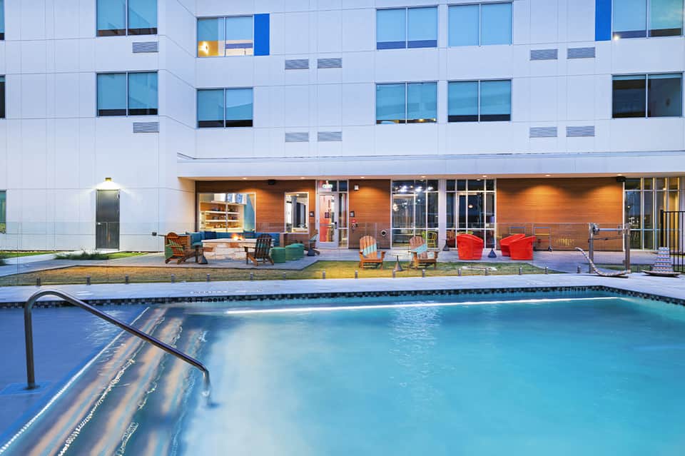 Modern hotel with white facade and pool area featuring colorful lounge chairs and seating