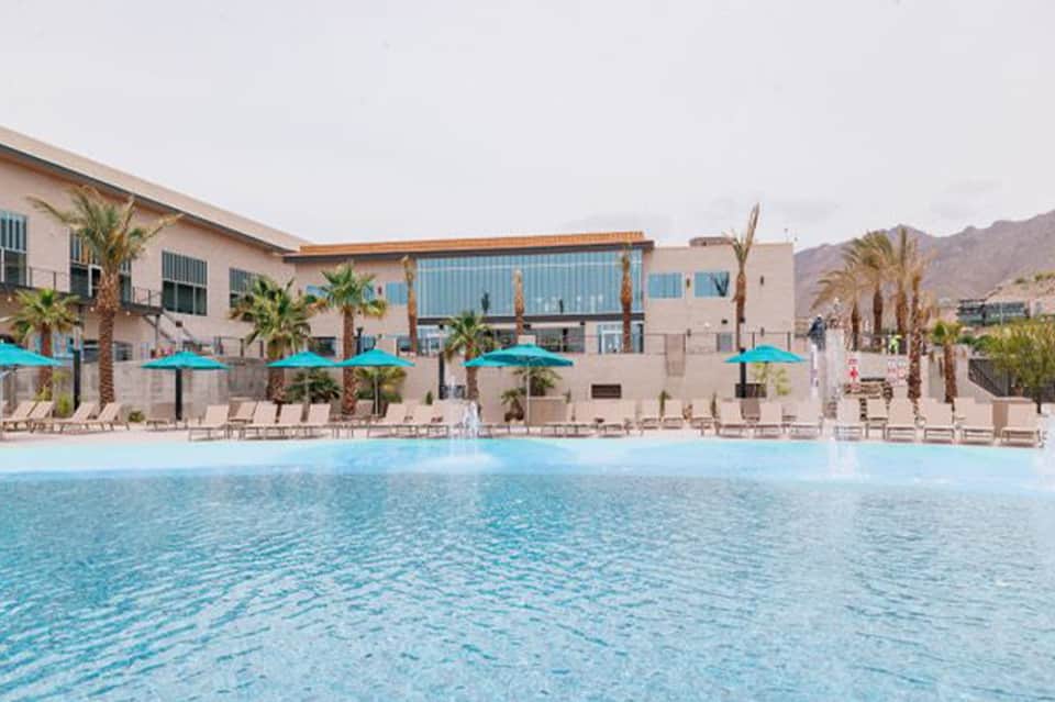 Resort pool with turquoise water, palm trees, blue umbrellas, and modern beige buildings