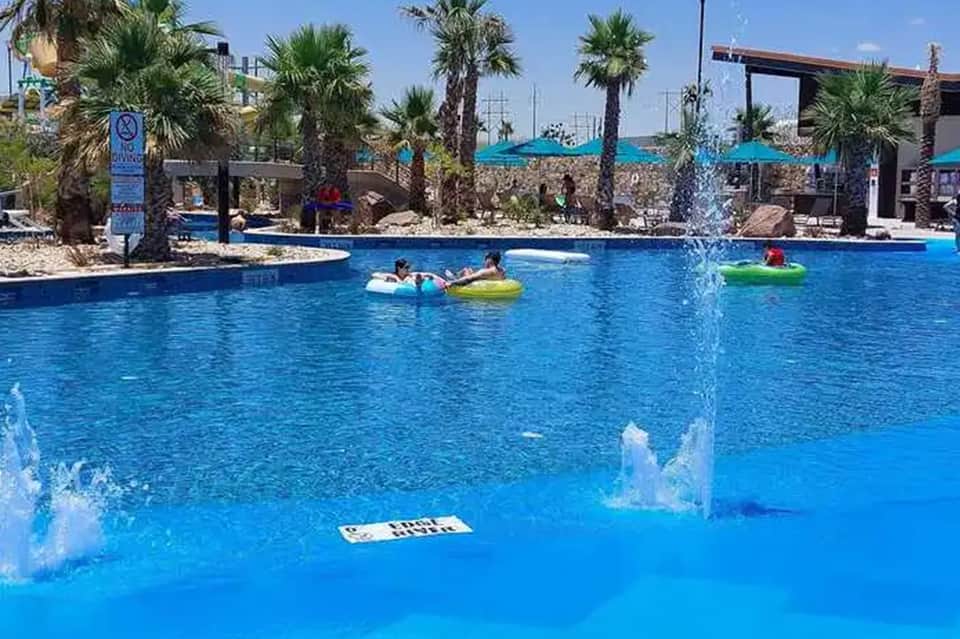 Resort pool with turquoise water, palm trees, blue umbrellas, and children playing
