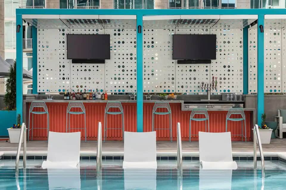 Poolside bar with turquoise frame, red corrugated metal, white bar stools, and pegboard backdrop