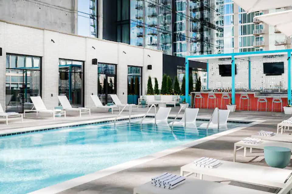 Contemporary rooftop pool with lounge chairs, turquoise pergola, red bar, and city skyline backdrop
