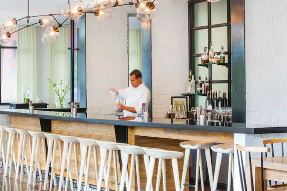 Contemporary bar with long counter, white stools, pendant lighting, and bartender in background